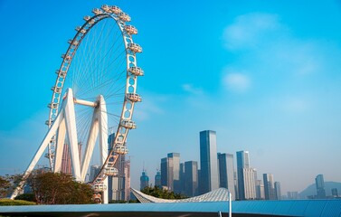 Fototapeta premium A large white ferris wheel against a blue sky with white clouds. The wheel is in front of a cityscape filled with tall buildings.