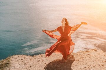 Woman red dress sea. Female dancer in a long red dress posing on a beach with rocks on sunny day. Girl on the nature on blue sky background.