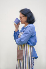 Side view: A woman wearing apron is having hot drink from a cup; one hand is folded