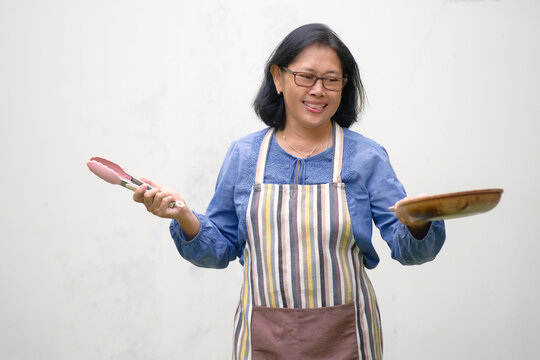 A woman wearing an apron holds a frying pan in one hand and food tongs in the other; smiling, happy expression