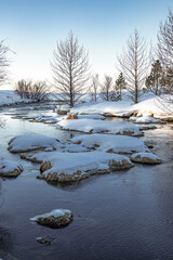 Wonderful morning winter view of a small Icelandic river with some trees and riverbanks covered with snow. Beautiful natural environment, peaceful background in Iceland. Snowy landscape with river.