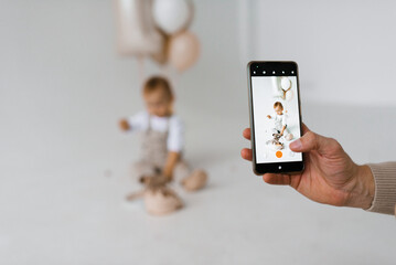 Father takes pictures of his son on his smartphone. Baby boy is sitting on the floor and eating a birthday cake