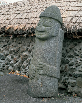 A Dolhareubang Stone is standing besides wall of a thatched house at Seongeup Folk Village near Seogwipo-si, Jeju-do, South Korea