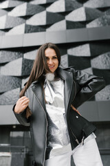 Hipster woman in a black jacket and white pants stands in front of a building