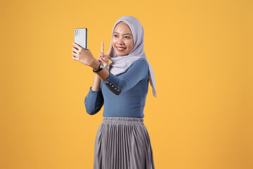 happy asian indonesian muslim woman holding smart phone giving victory finger gesture on isolated yellow background