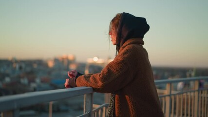 A sorrowful woman walks slowly while holding onto a metal railing, dressed in a brown coat and a hoodie, with a melancholic expression as the cityscape blurs in the background during sunset