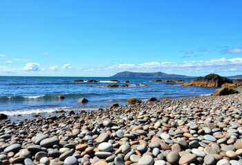A pebbles beach, shoreline with rocks and water and a bright blue sky.
