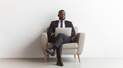 Man in a suit and tie, wearing black leather shoes, sitting on a small single-person sofa, laptop on his lap.