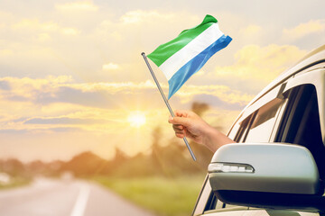 Waving the Sierra Leone flag against the sunrise or sunset from a car driving along a country road. Holding the Sierra Leone flag, traveling by car, on a weekend trip.