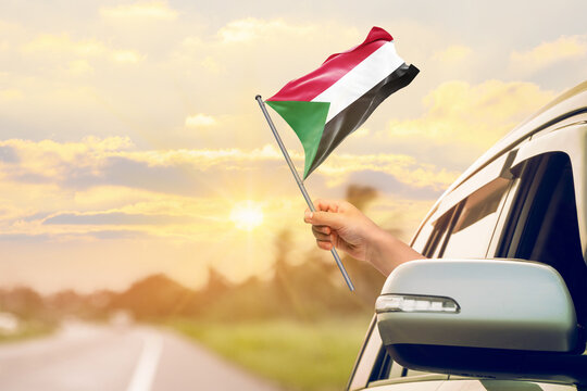 Waving the Sudan flag against the sunrise or sunset from a car driving along a country road. Holding the Sudan flag, traveling by car, on a weekend trip.