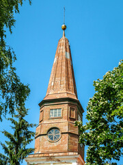 A tall, brown tower with a clock on the front framed by green trees and blue clear sky in the...