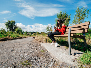 A teenager girl is sitting on a bench in a park, looking at her phone. The sky is blue and there are trees in the background and her small Yorkshire terrier is walking in the background. Sunny day.