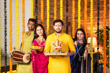 Indian couple playing dholak and cymbals, joyfully welcoming Ganesh idol with festive cheer at home