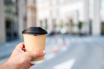 Close-up of hand holding takeaway coffee cup with a black lid, blurred city street background....