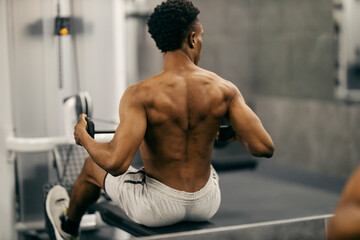 Rear view of strong muscular diverse bodybuilder practicing on cable machine at gym.