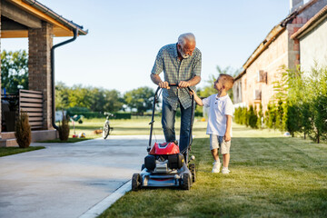 Front view of granddad and grandson mowing lawn in backyard together.