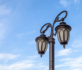 Black lamppost with three lamps and patterns on the background of blue sky with clouds