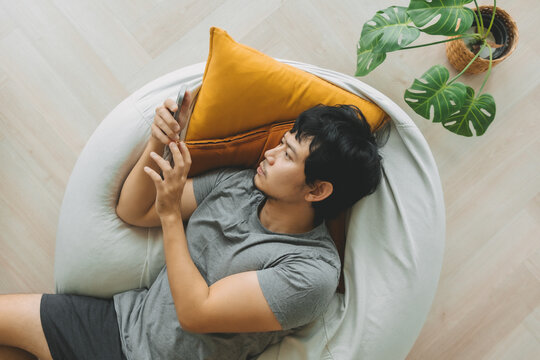 Asian man lean on the bean bag sofa using smartphone in the living room.