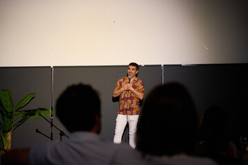 Male performer delivering a monologue on stage in a dimly lit theater with audience