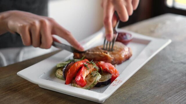 Girl cuts a cooked pork steak served on a white plate with grilled vegetables. Young woman using knife and fork to cutting meat eating in a restaurant. Balanced diet, cooking, culinary. Food concept