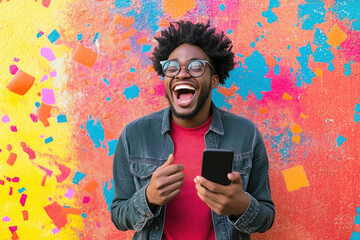 Happy young man laughing while holding smartphone against colorful graffiti wall, expressing joy and excitement, wearing glasses, denim jacket
