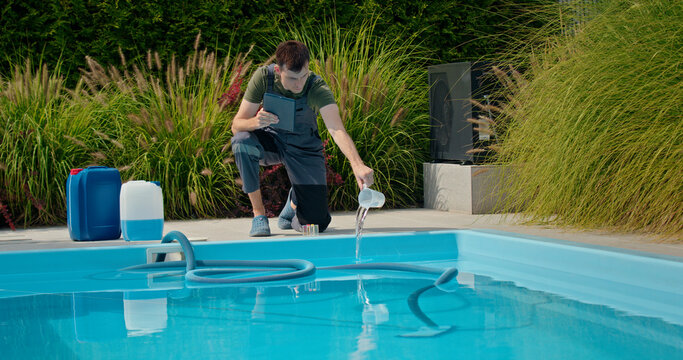 Pool technician pouring liquid into the pool while using a tablet, with chemical containers and greenery in the background.