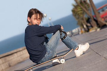 preteen boy with cool attitude sitting on skateboard smiling and looking at camera with street and...