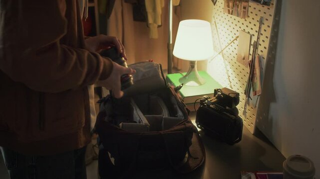 Side tilt shot of young Caucasian guy in casual clothes packing video camera and lens into bag before leaving home