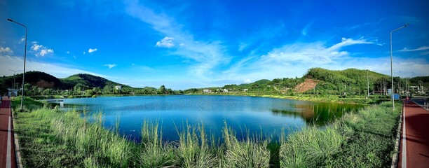 A serene lake with clear blue waters, surrounded by lush greenery and hills under a bright blue sky with few clouds. The tranquil setting is picturesque, ideal for nature lovers and outdoor activities