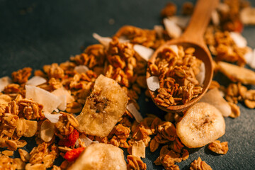 Overhead view of a wooden spoon with scattered granola, dried banana chips, and coconut flakes on a dark textured background