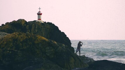 Man standing on the rocks setting up a fishing rod with a church cross on the mountain peak shot at Vagator beach goa showing the leisure and tourist activities
