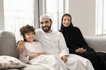 Obraz premium Positive Arabian mom and dad relaxing on comfortable home couch, embracing cheerful little son boy, looking at camera, smiling, laughing, having fun, enjoying family leisure, close relationship