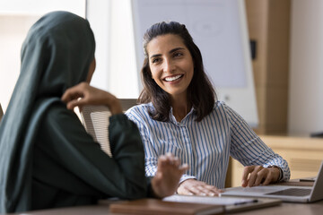 Positive Hispanic business professional woman talking to Arab female coworker in hijab, discussing...