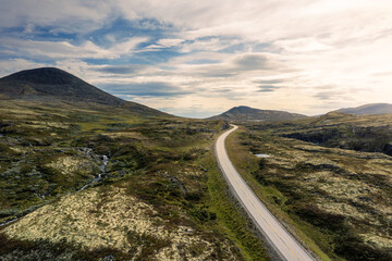 Bright sunlight illuminates the Muen and Nørdre Bølhøgda mountains as a winding road and mountain stream flow through the lush green landscape of Rondane National Park in Norway