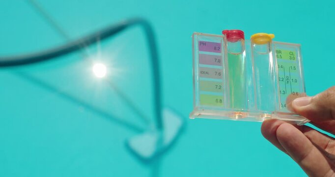 A hand holds a water test kit over a clear blue swimming pool with sunlight reflecting off the water.
