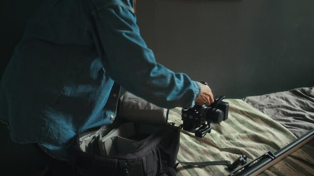 Tilt shot of young Caucasian lady in denim jacket and cap packing video camera and lens into bag before leaving home