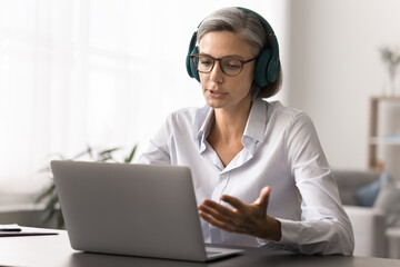 Middle-aged woman engaged in videoconference call, wearing wireless headphones, working in office, sit at desk with laptop, speaking, explaining information to client, engaged in virtual meeting event