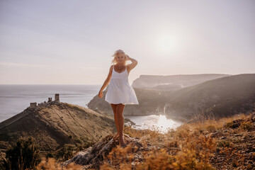 A blonde woman stands on a hill overlooking the ocean. She is wearing a white dress and she is enjoying the view.