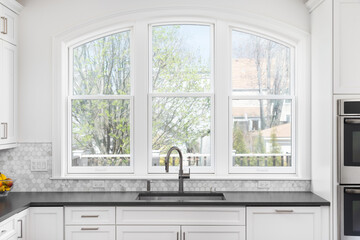 A kitchen detail with large arched windows, white cabinets, a dark grey countertop, stainless steel...