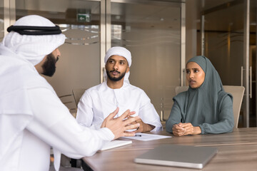 Male and female Arabian business partners in Muslim headwear and dresses talking at large office table, discussing solutions for project, cooperation, teamwork, company investment