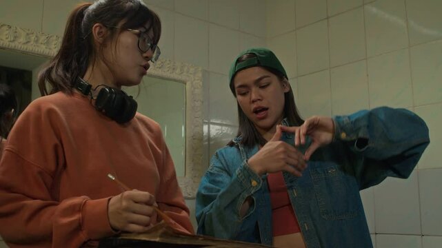 Medium shot of Caucasian and Asian ladies from film college planning interior of movie set using storyboard in washroom