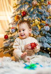 Child joyfully decorating a Christmas tree with a red ornament indoors. A cheerful child sits on the floor, holding a red ornament while surrounded by a beautifully decorated Christmas tree.