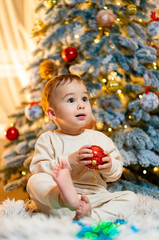 A young child sits happily by a decorated Christmas tree at home. A joyful toddler holds a red ornament while surrounded by a festive, decorated Christmas tree