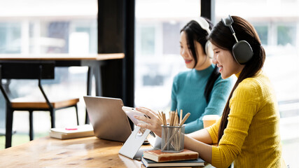 Two woman students wearing headphone studying at the university are preparing for the exam in the library on a laptop, discussing a studying