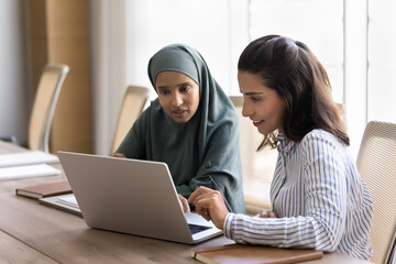 Two focused young Arab and Hispanic managers women working at laptop together, watching online work...