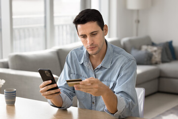 Focused young Latin consumer man making money transaction on online bank financial application, using cellphone and plastic credit card, paying for purchase on webstores, inserting data