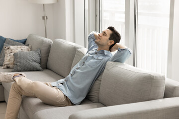 Obraz premium Calm handsome Latin man relaxing on comfortable couch at cozy home, leaning on soft back, breathing fresh cool, sitting air with closed eyes and open arms, enjoying on work break, comfort, leisure
