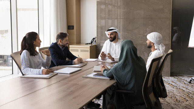 Happy young Arab business leader man talking to diverse partners at meeting. Multiethnic business team of Arabic and European colleagues discussing cooperation, teamwork. Banner shot
