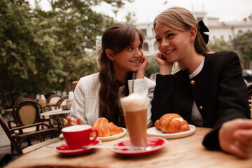 Two beautiful young girls are sitting in a French cafe, eating a croissant and drinking coffee with whipped cream. Teenagers are dressed in business style, black and white jacket in Old Money style