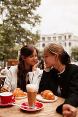 Two beautiful young girls are sitting in a French cafe, eating a croissant and drinking coffee with whipped cream. Teenagers are dressed in business style, black and white jacket in Old Money style